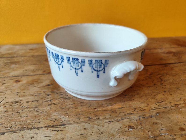 K&ouml;nnte beinhalten: Eine wei&szlig;e Keramiksch&uuml;ssel mit einem blauen und wei&szlig;en Blumenmuster am Rand. Die Sch&uuml;ssel hat zwei kleine Griffe an den Seiten.
