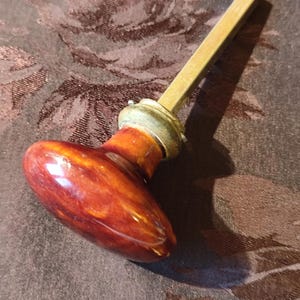 Op de afbeelding: Een vintage deurknop met een gepolijste, roodbruine ovale handgreep. De handgreep is verbonden met een messingkleurige vierkante staaf en een decoratieve metalen kraag. De knop heeft een glanzende afwerking, wat suggereert dat hij van hout of een vergelijkbaar materiaal is.