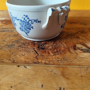 Op de afbeelding: Witte keramische soepkom met een blauw bloemenmotief en een enkele handgreep. De kom staat op een houten oppervlak.