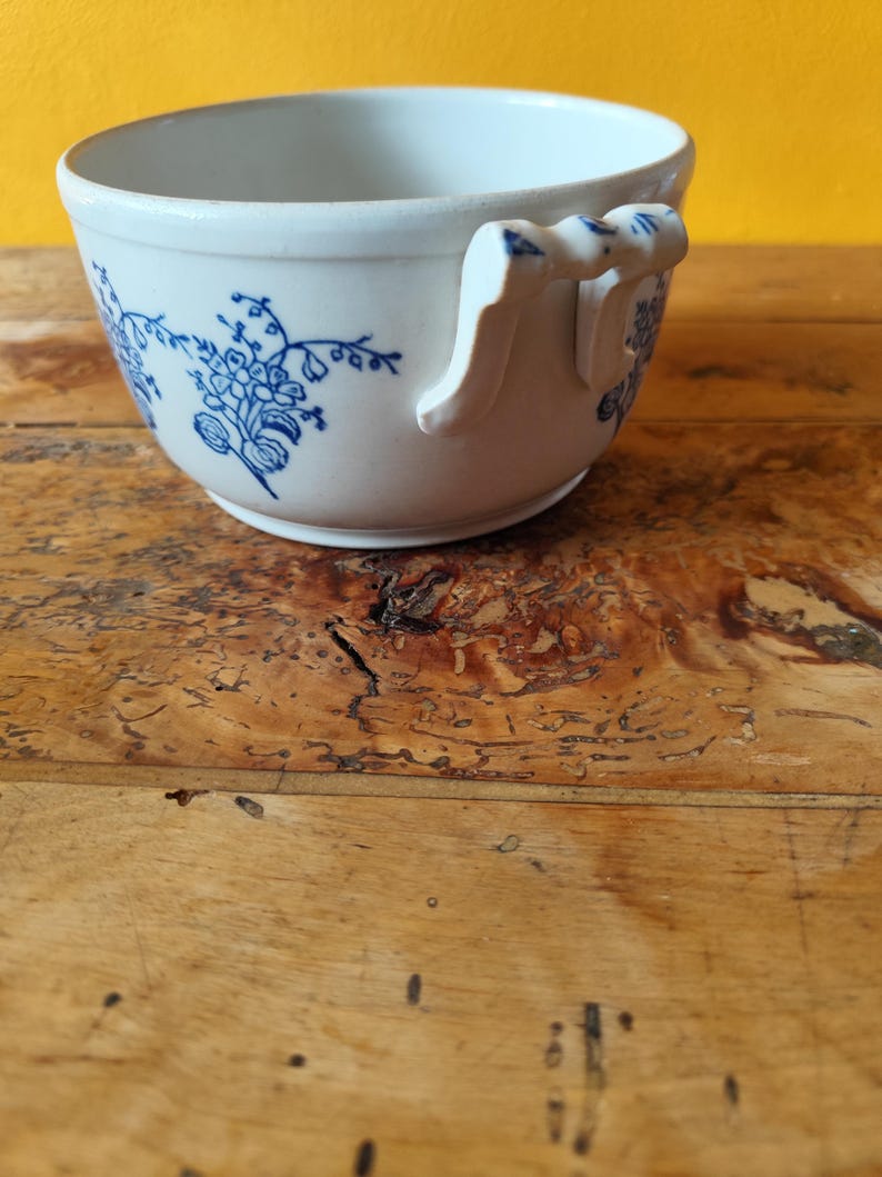 Op de afbeelding: Witte keramische soepkom met een blauw bloemenmotief en een enkele handgreep. De kom staat op een houten oppervlak.