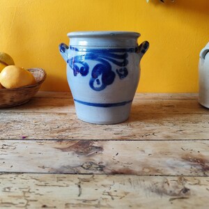 Może przedstawiać: Jasnoniebieski ceramiczny garnek z dwoma małymi uchwytami i niebieskim kwiatowym wzorem. Garnek ma lekko rozszerzający się kształt i stoi na drewnianej powierzchni. Tło jest w jednolitym żółtym kolorze.
