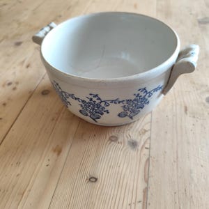 Op de afbeelding: Een witte keramische kom met blauwe bloemenmotieven en twee handvatten. De kom staat op een houten oppervlak.