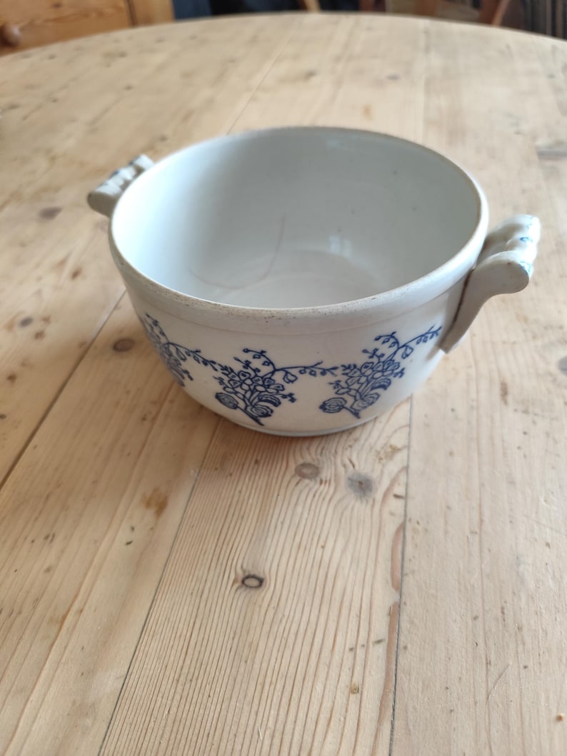 Op de afbeelding: Een witte keramische kom met blauwe bloemenmotieven en twee handvatten. De kom staat op een houten oppervlak.