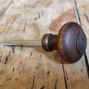 May include: A vintage key with a dark brown, wooden handle and a brass-coloured blade. The handle is round with a circular design on the end. The key is lying on a wooden surface.