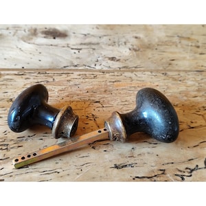 May include: Two antique black door knobs with brass accents and a connecting rod, resting on a weathered wooden surface. The knobs have an oval shape and a glossy finish, showcasing a vintage aesthetic.