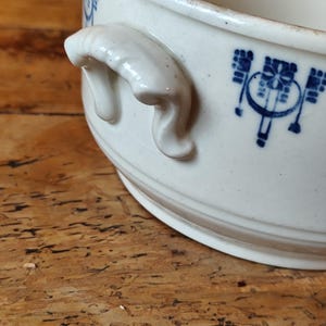 K&ouml;nnte beinhalten: Wei&szlig;e Keramiksch&uuml;ssel mit blau-wei&szlig;em Blumendesign und zwei Griffen. Die Sch&uuml;ssel steht auf einer Holzoberfl&auml;che.