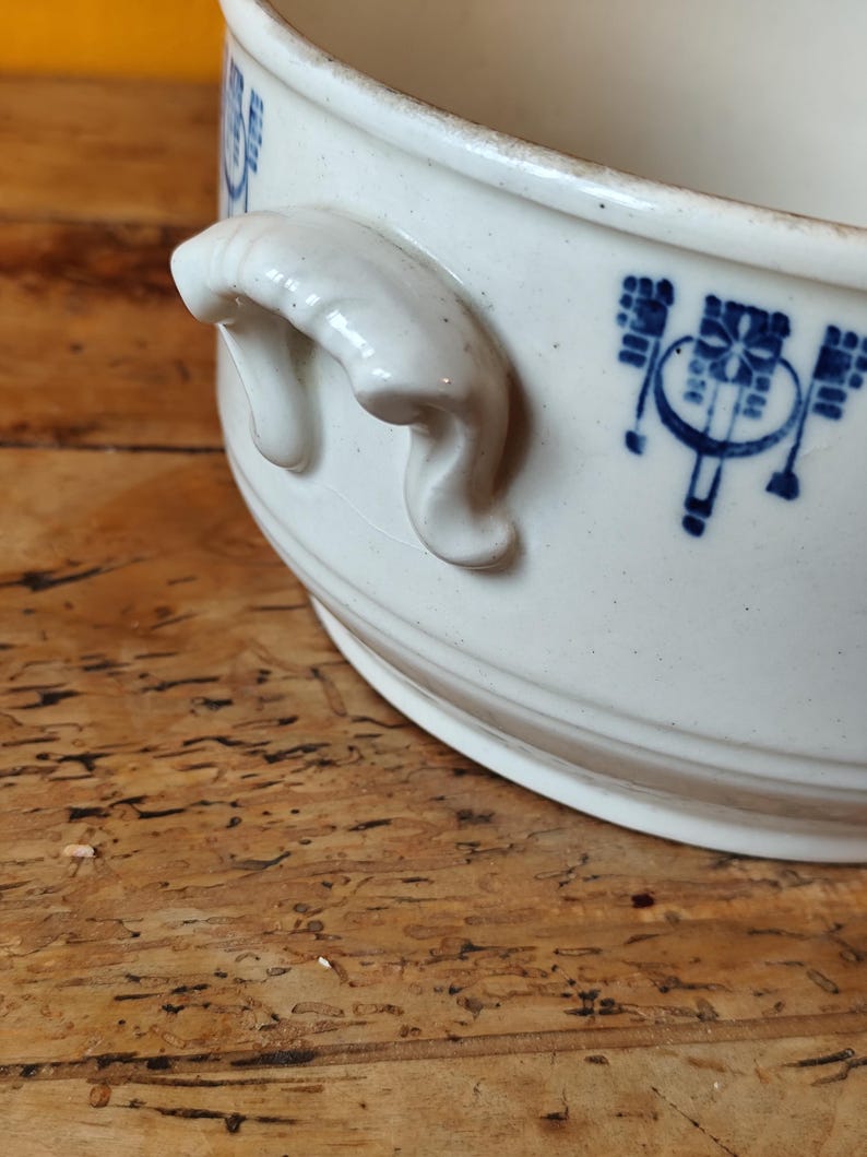 K&ouml;nnte beinhalten: Wei&szlig;e Keramiksch&uuml;ssel mit blau-wei&szlig;em Blumendesign und zwei Griffen. Die Sch&uuml;ssel steht auf einer Holzoberfl&auml;che.