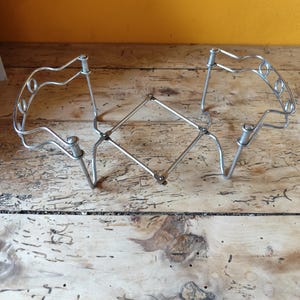 May include: A silver-coloured metal expandable trivet featuring a central diamond-shaped lattice mechanism connected to two curved, raised side supports. The structure is constructed from thin wire rods and includes small circular loops along the top edges.
