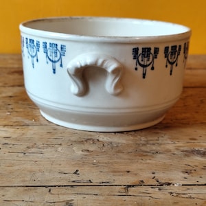 K&ouml;nnte beinhalten: Wei&szlig;e Keramiksch&uuml;ssel mit blau-wei&szlig;em Blumendesign und zwei Griffen. Die Sch&uuml;ssel steht auf einer Holzoberfl&auml;che.