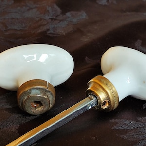 May include: Two antique white porcelain door knobs with brass accents. One knob is detached, revealing the metal rod. The knobs have an oval shape and are set against a dark, textured background.