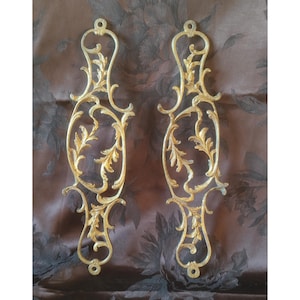 May include: Two decorative gold-coloured metal pieces with an ornate design. Each piece has a symmetrical pattern of scrolling leaves and floral details. They have a curved shape with loops at the top and bottom for hanging or fixing.