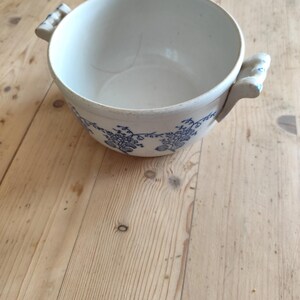 Op de afbeelding: Een witte keramische kom met blauwe bloemenmotieven en twee handvatten. De kom staat op een houten oppervlak.