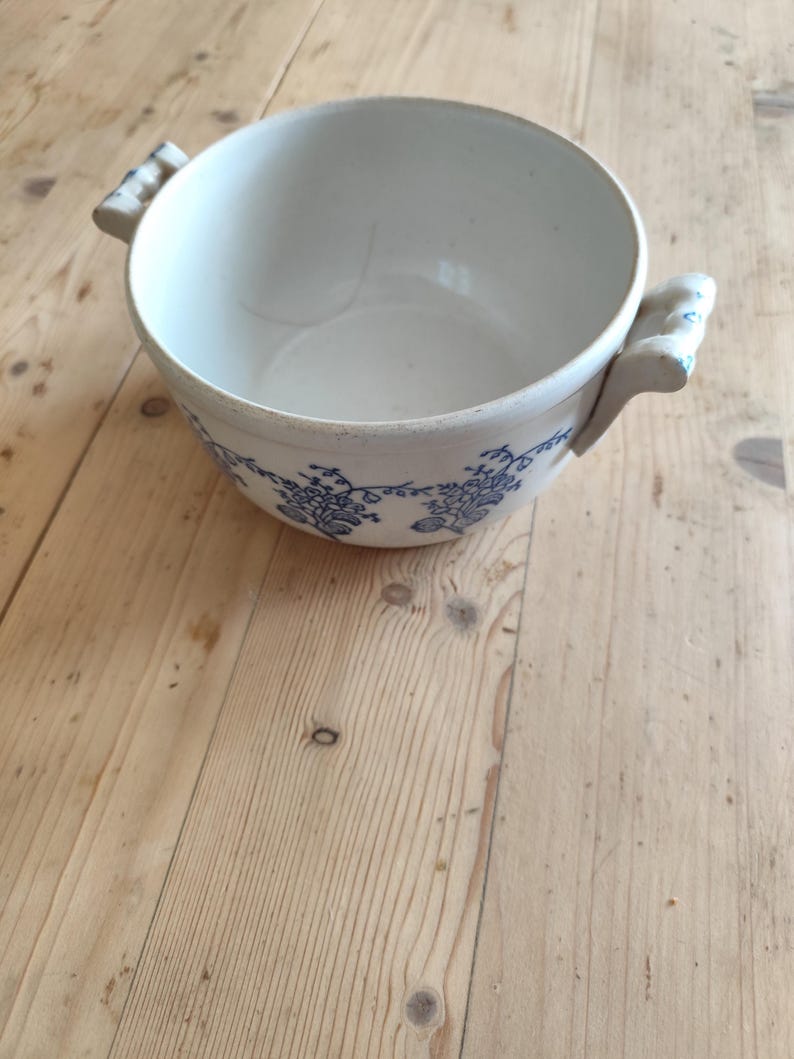 Op de afbeelding: Een witte keramische kom met blauwe bloemenmotieven en twee handvatten. De kom staat op een houten oppervlak.