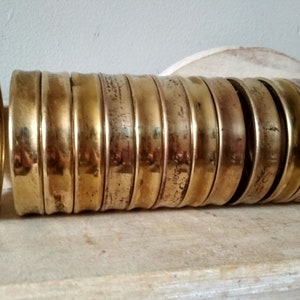 May include: A stack of 15 brass rings, each with a slightly different width and a smooth, polished finish. The rings are stacked on a wooden surface.