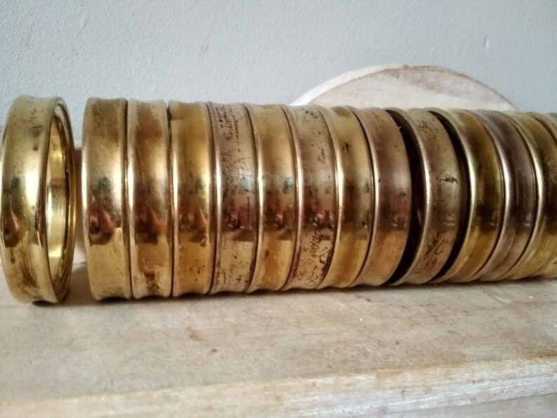 May include: A stack of 15 brass rings, each with a slightly different width and a smooth, polished finish. The rings are stacked on a wooden surface.