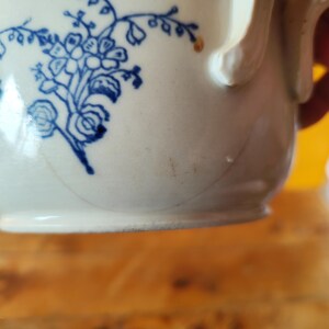 Op de afbeelding: Een witte keramische mok met een blauw bloemenmotief. De mok heeft een handvat en een licht afgeronde vorm.