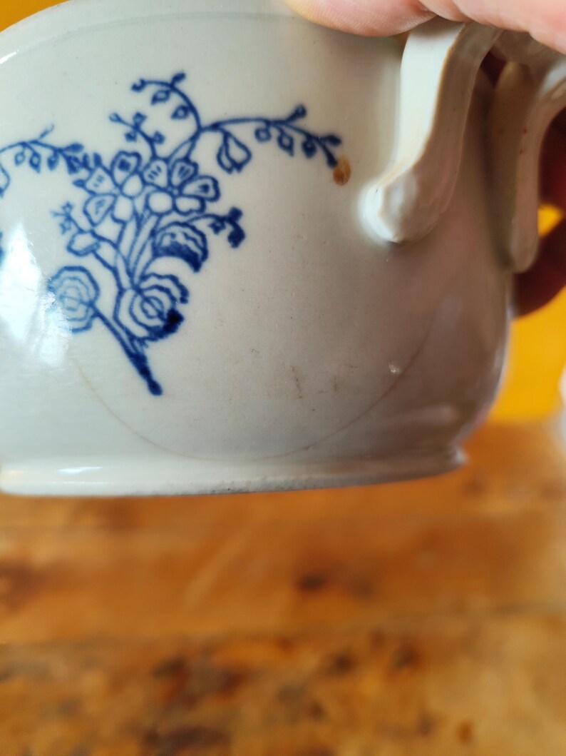 Op de afbeelding: Een witte keramische mok met een blauw bloemenmotief. De mok heeft een handvat en een licht afgeronde vorm.