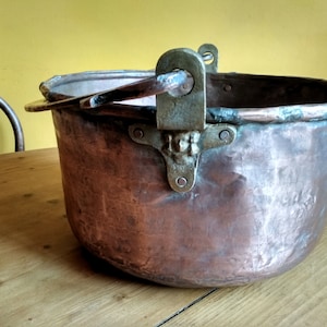 Peut inclure: Une grande marmite en cuivre antique avec une poignée en laiton et une tête de lion décorative en laiton sur le côté. La marmite a une patine usée et vieillie et repose sur une surface en bois.