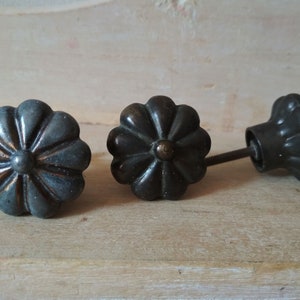 May include: Three dark grey metal cabinet knobs with a flower design. The knobs have a slightly raised centre and a slightly raised edge.