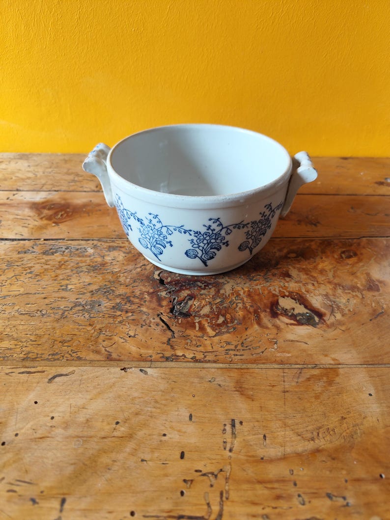 Op de afbeelding: Een witte keramische kom met een blauw bloemenpatroon en twee handvatten. De kom staat op een houten oppervlak.