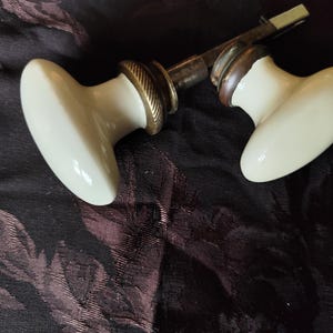 Puede incluir: Dos pomos de puerta antiguos con un diseño de porcelana blanca crema de forma ovalada. Los pomos tienen una base de color latón y una varilla metálica que los conecta. La ferretería es probablemente para una puerta vintage.