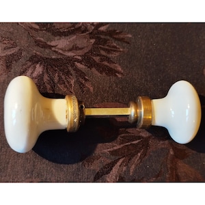 May include: A vintage door knob set featuring two white porcelain knobs connected by a brass rod. The knobs have a rounded, oval shape and are accented with brass collars. The hardware is likely for a door or cabinet.
