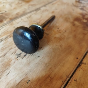 Peut inclure: Un bouton de porte noir de forme ovale, avec une collerette en laiton et une tige en métal sombre. Le bouton présente des signes d'usure. Le fond est une surface en bois avec un grain naturel.