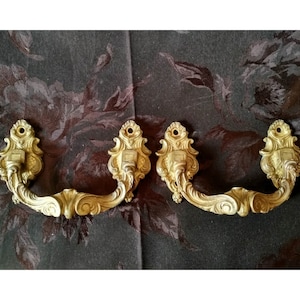 Pode incluir: Duas puxadores de gaveta de metal dourado ornamentado com um design de volutas decorativas. As puxadores têm uma abertura em forma de D e são projetadas para serem montadas em um armário ou uma cômoda.