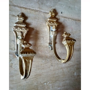 Può includere: Due ganci in metallo dorato decorati con un designo decorativo. I ganci sono montati su una superficie in legno.