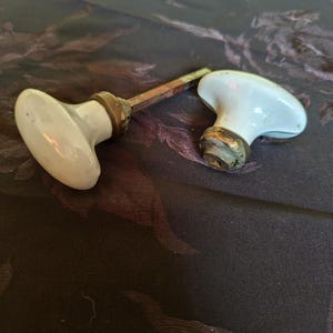 May include: Two antique white porcelain door knobs with brass accents. The knobs are on a dark fabric background.