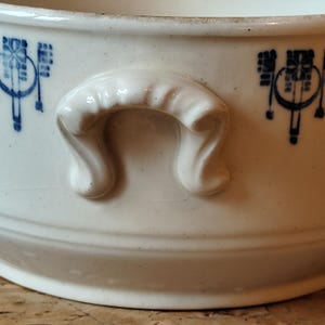 K&ouml;nnte beinhalten: Wei&szlig;e Keramiksch&uuml;ssel mit blau-wei&szlig;em Blumenmuster und zwei Griffen. Die Sch&uuml;ssel ist oval geformt und hat einen leicht erh&ouml;hten Rand.