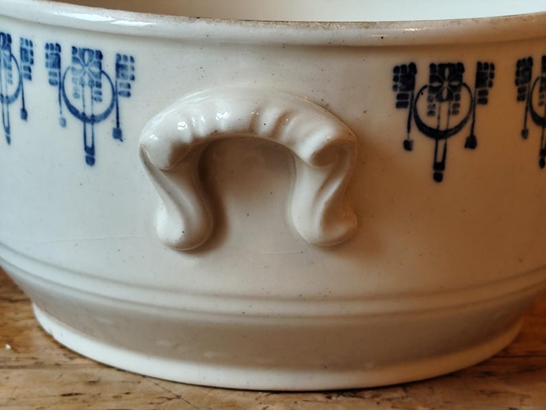 K&ouml;nnte beinhalten: Wei&szlig;e Keramiksch&uuml;ssel mit blau-wei&szlig;em Blumenmuster und zwei Griffen. Die Sch&uuml;ssel ist oval geformt und hat einen leicht erh&ouml;hten Rand.