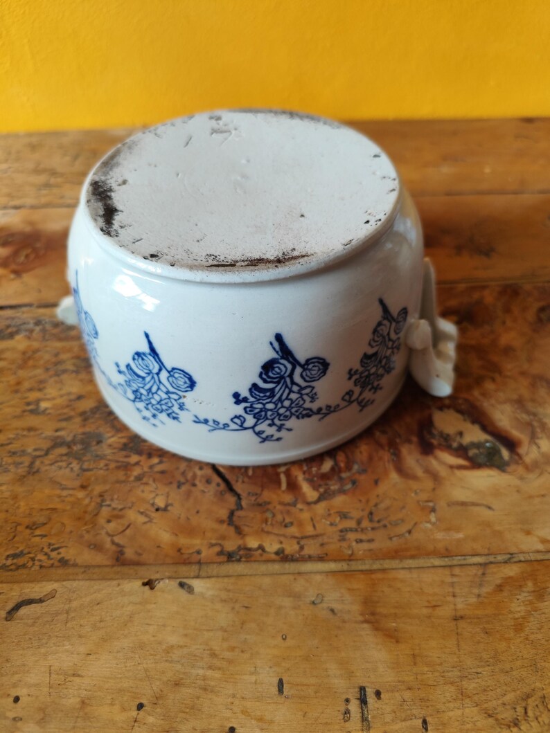 Op de afbeelding: Witte keramische pot met een blauw bloemenontwerp en twee handvatten. De pot staat op een houten oppervlak.