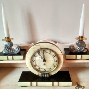 May include: A vintage Art Deco style clock with two candle holders. The clock face is white with black Roman numerals and black hands. The clock is made of marble and has a black base. The candle holders are made of silver and have a gold base.