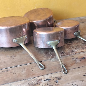 May include: Four copper pots with brass handles rest on a wooden surface. The pots vary in size, with rounded bodies and straight sides. The copper has a warm, reddish-brown hue, and the brass handles are attached with rivets.