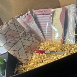 May include: A gift box filled with stationery supplies, including a pink and gold marble-patterned notebook, a pack of pink and white striped sticky notes, a pack of yellow and pink sticky notes with the text "Special moments", a clear plastic bag of white stickers, and a pink highlighter.