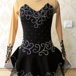 Puede incluir: Vestido negro de patinaje art&iacute;stico con escote coraz&oacute;n y mangas largas color nude. El corpi&ntilde;o y la falda est&aacute;n adornados con pedrer&iacute;a blanca en un patr&oacute;n floral. Una superposici&oacute;n de falda negra transparente a&ntilde;ade volumen.