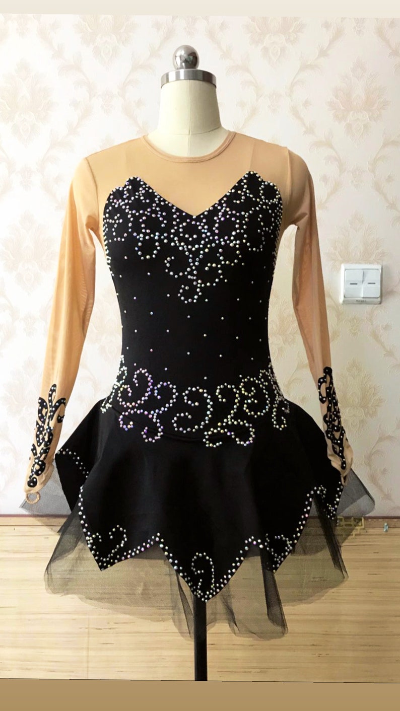 Puede incluir: Vestido negro de patinaje art&iacute;stico con escote coraz&oacute;n y mangas largas color nude. El corpi&ntilde;o y la falda est&aacute;n adornados con pedrer&iacute;a blanca en un patr&oacute;n floral. Una superposici&oacute;n de falda negra transparente a&ntilde;ade volumen.