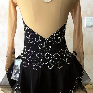 Puede incluir: Vestido negro de patinaje art&iacute;stico con espalda y mangas de malla color nude. El corpi&ntilde;o est&aacute; adornado con un patr&oacute;n arremolinado de pedrer&iacute;a iridiscente. La falda tiene un dobladillo de tul en capas. El vestido se exhibe en un maniqu&iacute;.