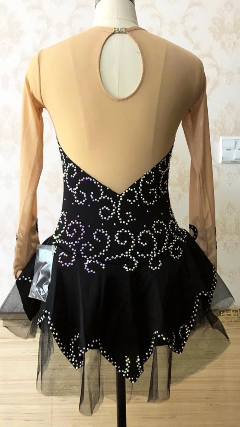 Puede incluir: Vestido negro de patinaje art&iacute;stico con espalda y mangas de malla color nude. El corpi&ntilde;o est&aacute; adornado con un patr&oacute;n arremolinado de pedrer&iacute;a iridiscente. La falda tiene un dobladillo de tul en capas. El vestido se exhibe en un maniqu&iacute;.