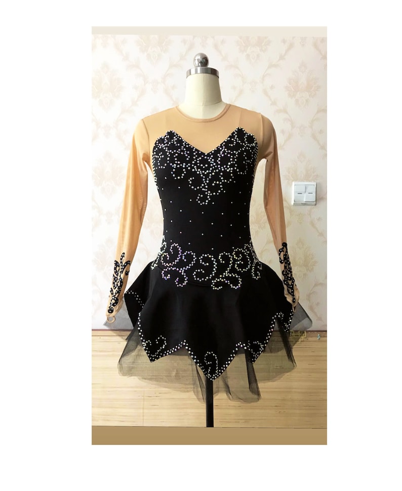 Puede incluir: Vestido negro de patinaje art&iacute;stico con mangas largas color nude y falda negra. El corpi&ntilde;o est&aacute; adornado con pedrer&iacute;a blanca. La falda tiene un dobladillo con volantes y una enagua negra transparente.