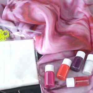 May include: A tie-dye silk scarf in shades of pink and red, with a kit containing dye bottles, rubber bands, and a plastic bag. The dye bottles are in various colors, including pink, orange, purple, and white.