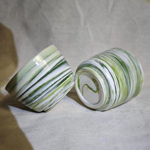 May include: Two ceramic bowls with a green and white marbled design. The bowls are sitting on a white fabric surface.