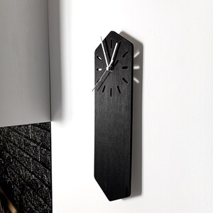 May include: A vertical, elongated, hexagonal black wall clock with a flat, matte finish. The clock face features thin, silver-coloured hour and minute hands, with simple, laser-cut slits serving as hour markers arranged in a circular pattern.