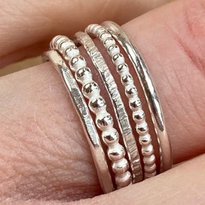 May include: Five silver rings of varying designs are stacked on a finger. The rings include smooth bands, beaded, textured, and hammered styles. The image highlights the different textures and designs of the rings, showcasing their individual characteristics.