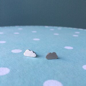 May include: Two cloud-shaped stud earrings. One is white, the other grey. The earrings are small and delicate. They are displayed on a light blue polka-dot surface. The earrings are ideal for everyday wear.