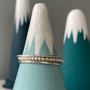 May include: Three silver rings are stacked on a light blue cone-shaped ring holder. The ring holder is in front of two other similar holders, painted with a mountain design in white and teal.