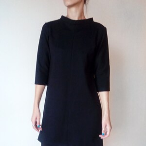 May include: A black shift dress with a mock neck and three-quarter sleeves. The dress is knee-length and has a straight silhouette. The model is wearing black tights and black ankle boots.