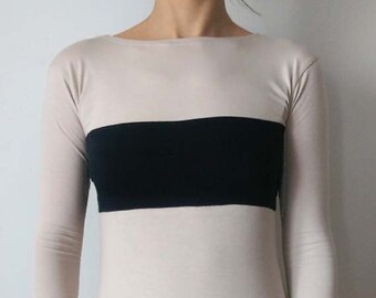 May include: A long-sleeved shirt with a black band across the chest. The shirt is beige and the band is black. The shirt is being worn by a person.