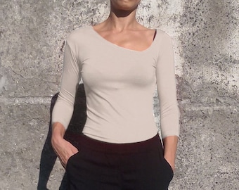 May include: A beige long-sleeved shirt with a scoop neck and a slightly asymmetrical neckline. The shirt is fitted and has a smooth, stretchy fabric. The model is wearing black pants.